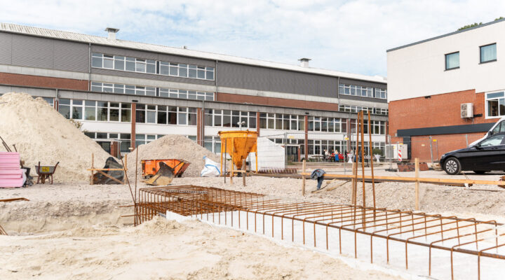 Baustelle vor einem größeren Gebäudekomplex. Im Vordergrund sind Sandflächen, Baugruben und Stahlbewehrungen für Fundamente zu sehen. Dahinter befinden sich Sandhaufen, Baugeräte und Absperrungen; im Hintergrund erstreckt sich ein mehrgeschossiges Gebäude.