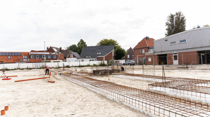 Weitläufige Baustelle mit vorbereiteten Fundamenten und zahlreichen Stahlbewehrungen im sandigen Untergrund. Zwei Bauarbeiter sind auf dem Gelände tätig. Im Hintergrund stehen Wohnhäuser sowie ein größeres Gewerbegebäude.