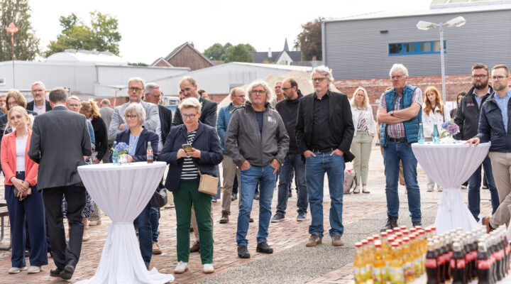 Eine größere Gruppe von Menschen steht bei einer Veranstaltung im Freien auf einem Baustellengelände. Einige Personen stehen an weißen Stehtischen mit Getränken und Blumen; im Vordergrund sind zahlreiche Getränkeflaschen aufgebaut. Im Hintergrund befinden sich Gewerbegebäude und Baumaterialien.
