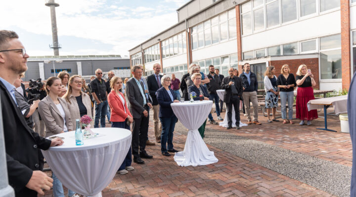 Eine größere Gruppe von Menschen verfolgt eine Veranstaltung im Freien vor einem mehrgeschossigen Gebäude. Die Gäste stehen an weiß gedeckten Stehtischen mit Blumen und Getränken; einige fotografieren oder filmen die Veranstaltung. Im Hintergrund sind weitere Gebäude und ein hoher Turm zu sehen.