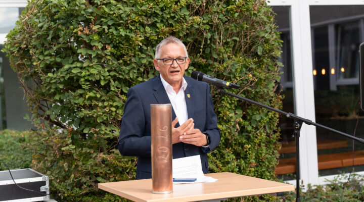 Landrat Olaf Meinen hält bei einer Veranstaltung im Freien eine Rede an einem Mikrofon. Vor ihm steht ein Tisch mit einer kupferfarbenen Zeitkapsel sowie Unterlagen. Im Hintergrund befindet sich eine große grüne Hecke.