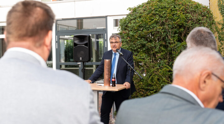 Schulleiter Volker Cammans hält bei einer Veranstaltung im Freien eine Rede an einem Mikrofon. Vor ihm steht ein Tisch mit einer kupferfarbenen Zeitkapsel und Unterlagen. Im Vordergrund sind mehrere Gäste von hinten zu sehen.
