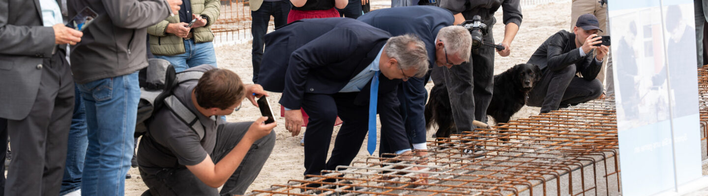 Mehrere Personen stehen auf einer Baustelle rund um eine vorbereitete Fundamentfläche mit Stahlbewehrung. Zwei Männer beugen sich über die Konstruktion und führen symbolisch Arbeiten daran aus, während Umstehende den Moment mit Smartphones und Kameras festhalten. Im Hintergrund sind Baugeräte, Absperrungen und Wohnhäuser zu sehen.