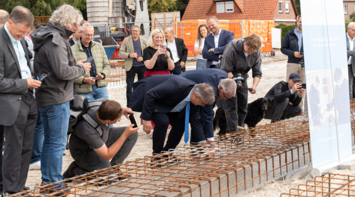 Mehrere Personen stehen auf einer Baustelle rund um eine vorbereitete Fundamentfläche mit Stahlbewehrung. Zwei Männer beugen sich über die Konstruktion und führen symbolisch Arbeiten daran aus, während Umstehende den Moment mit Smartphones und Kameras festhalten. Im Hintergrund sind Baugeräte, Absperrungen und Wohnhäuser zu sehen.