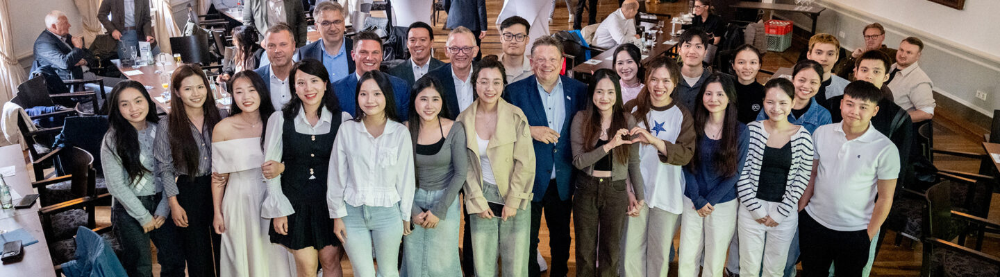 Große Gruppe junger Menschen und mehrerer Erwachsener posiert gemeinsam in einem festlichen Saal für ein Gruppenfoto. Im Hintergrund stehen weitere Gäste an Tischen und Stehtischen; der Raum ist mit hohen Fenstern, Kronleuchtern und Gemälden ausgestattet.