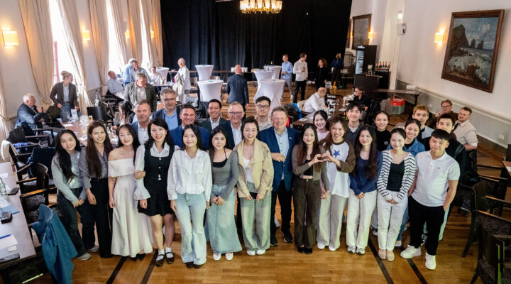Große Gruppe junger Menschen und mehrerer Erwachsener posiert gemeinsam in einem festlichen Saal für ein Gruppenfoto. Im Hintergrund stehen weitere Gäste an Tischen und Stehtischen; der Raum ist mit hohen Fenstern, Kronleuchtern und Gemälden ausgestattet.