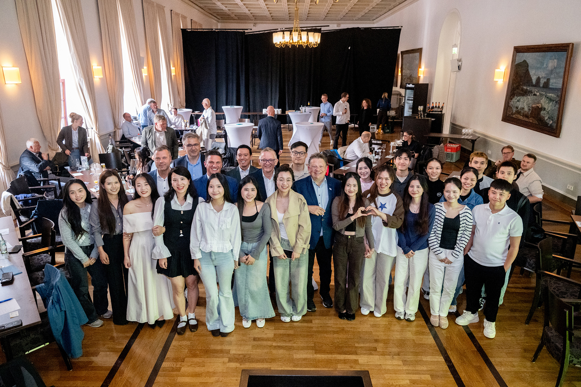 Große Gruppe junger Menschen und mehrerer Erwachsener posiert gemeinsam in einem festlichen Saal für ein Gruppenfoto. Im Hintergrund stehen weitere Gäste an Tischen und Stehtischen; der Raum ist mit hohen Fenstern, Kronleuchtern und Gemälden ausgestattet.