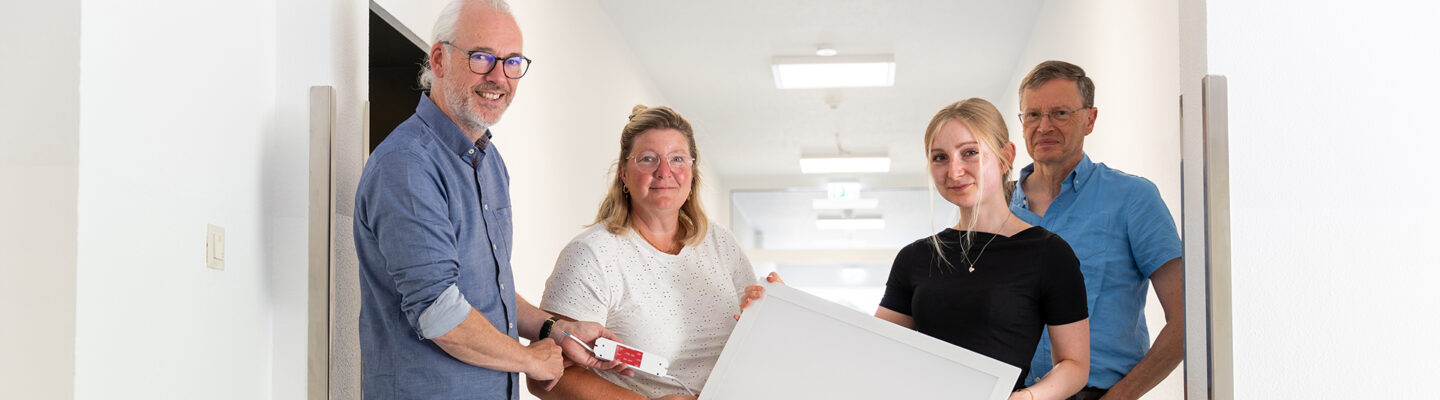 Vier Personen stehen in einem hellen Flur und blicken in die Kamera. Zwei von ihnen halten gemeinsam eine große quadratische LED-Deckenleuchte, eine weitere Person zeigt ein dazugehöriges Anschluss- oder Steuerungselement. Im Hintergrund sind mehrere bereits installierte Deckenleuchten zu sehen.