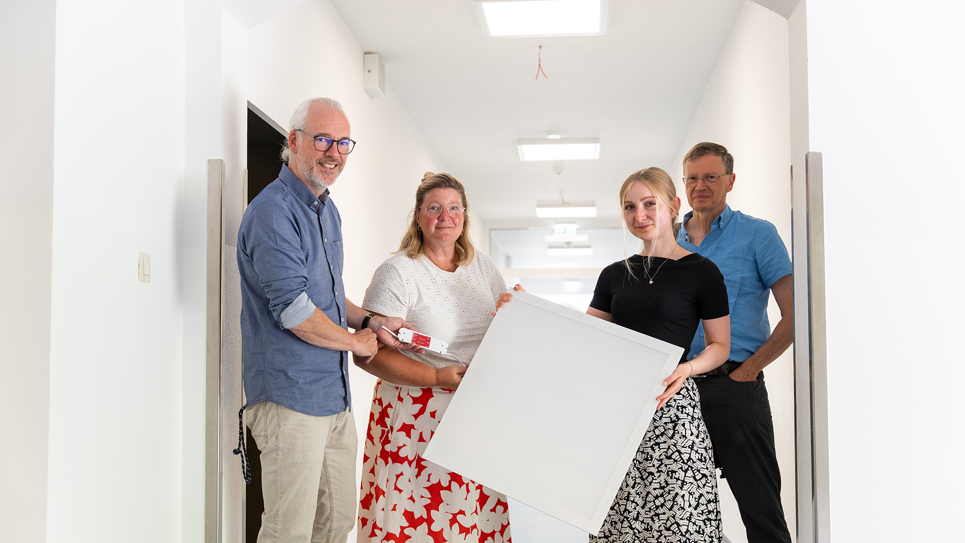 Vier Personen stehen in einem hellen Flur und blicken in die Kamera. Zwei von ihnen halten gemeinsam eine große quadratische LED-Deckenleuchte, eine weitere Person zeigt ein dazugehöriges Anschluss- oder Steuerungselement. Im Hintergrund sind mehrere bereits installierte Deckenleuchten zu sehen.