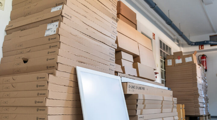 Mehrere Paletten mit flachen Kartons sind in einem Lagerraum gestapelt. Vor den Kartons lehnt eine große rechteckige LED-Leuchte. Weitere verpackte Leuchten und Materialien stehen im Hintergrund.