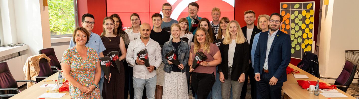 Gruppenfoto im Sitzungsraum des Landkreises Aurich: Rund 20 Auszubildende, Studierende und Mitarbeitende stehen gemeinsam vor einer großen roten Bildschirmwand mit der Aufschrift „Einführungstage für Auszubildende und Studierende – Herzlich Willkommen beim Landkreis Aurich!“. Einige der neuen Nachwuchskräfte halten schwarze Willkommensmappen mit dem Slogan „Deine Zukunft beginnt hier“ in den Händen. Rechts und links stehen Konferenztische mit Getränken, Unterlagen und roten Stoffbeuteln bereit. Die Gruppe lächelt in die Kamera und vermittelt einen freundlichen, offiziellen Auftakt für den Ausbildungs- und Studienstart beim Landkreis Aurich.