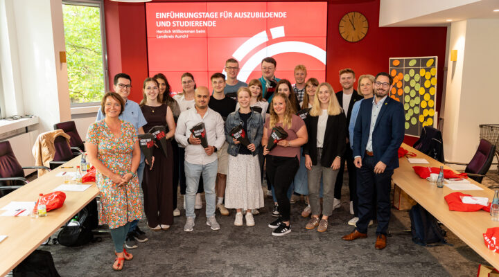 Gruppenfoto im Sitzungsraum des Landkreises Aurich: Rund 20 Auszubildende, Studierende und Mitarbeitende stehen gemeinsam vor einer großen roten Bildschirmwand mit der Aufschrift „Einführungstage für Auszubildende und Studierende – Herzlich Willkommen beim Landkreis Aurich!“. Einige der neuen Nachwuchskräfte halten schwarze Willkommensmappen mit dem Slogan „Deine Zukunft beginnt hier“ in den Händen. Rechts und links stehen Konferenztische mit Getränken, Unterlagen und roten Stoffbeuteln bereit. Die Gruppe lächelt in die Kamera und vermittelt einen freundlichen, offiziellen Auftakt für den Ausbildungs- und Studienstart beim Landkreis Aurich.