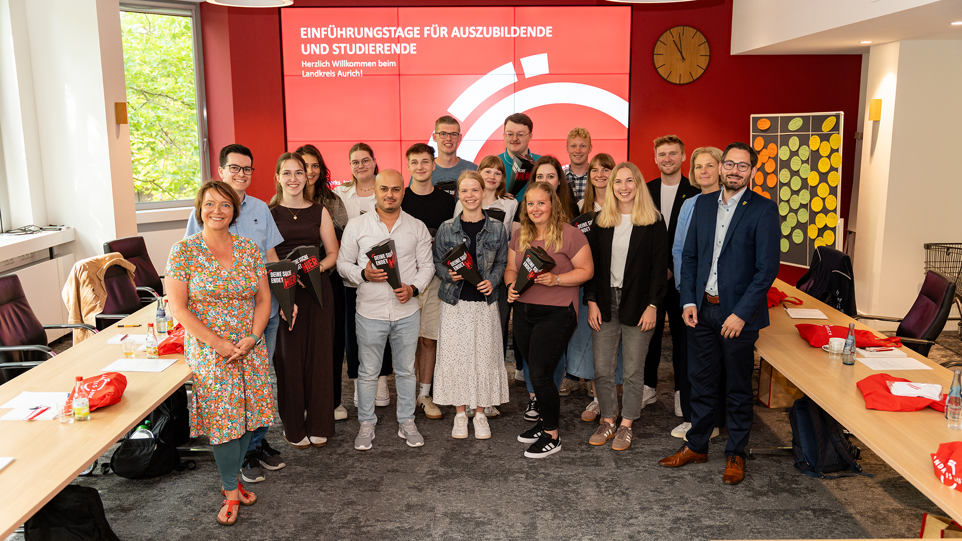 Gruppenfoto im Sitzungsraum des Landkreises Aurich: Rund 20 Auszubildende, Studierende und Mitarbeitende stehen gemeinsam vor einer großen roten Bildschirmwand mit der Aufschrift „Einführungstage für Auszubildende und Studierende – Herzlich Willkommen beim Landkreis Aurich!“. Einige der neuen Nachwuchskräfte halten schwarze Willkommensmappen mit dem Slogan „Deine Zukunft beginnt hier“ in den Händen. Rechts und links stehen Konferenztische mit Getränken, Unterlagen und roten Stoffbeuteln bereit. Die Gruppe lächelt in die Kamera und vermittelt einen freundlichen, offiziellen Auftakt für den Ausbildungs- und Studienstart beim Landkreis Aurich.