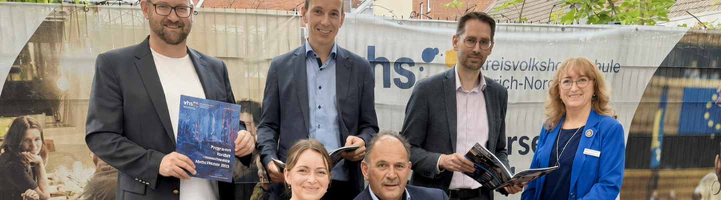 Gruppenfoto von sechs Personen vor einem Banner der Kreisvolkshochschule Aurich-Norden. Vier Männer und zwei Frauen stehen beziehungsweise sitzen um eine Holzbank und halten Programmhefte oder Broschüren in den Händen. Im Hintergrund sind das Logo und der Schriftzug der Kreisvolkshochschule zu sehen. Die Aufnahme entstand im Freien vor einer Kulisse mit Häusern und Grünpflanzen.