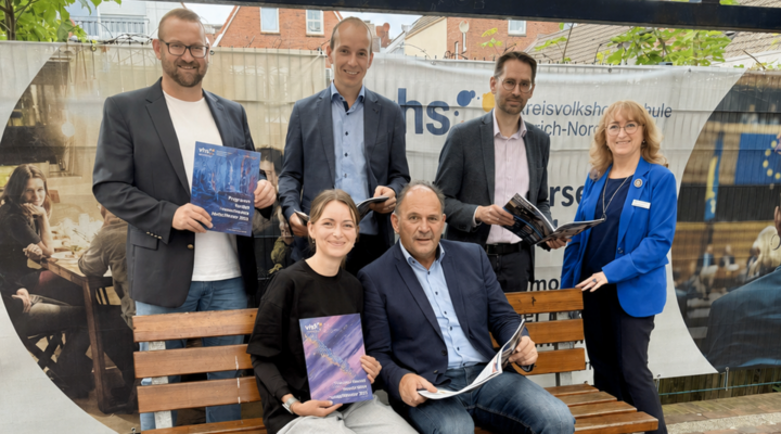 Gruppenfoto von sechs Personen vor einem Banner der Kreisvolkshochschule Aurich-Norden. Vier Männer und zwei Frauen stehen beziehungsweise sitzen um eine Holzbank und halten Programmhefte oder Broschüren in den Händen. Im Hintergrund sind das Logo und der Schriftzug der Kreisvolkshochschule zu sehen. Die Aufnahme entstand im Freien vor einer Kulisse mit Häusern und Grünpflanzen.
