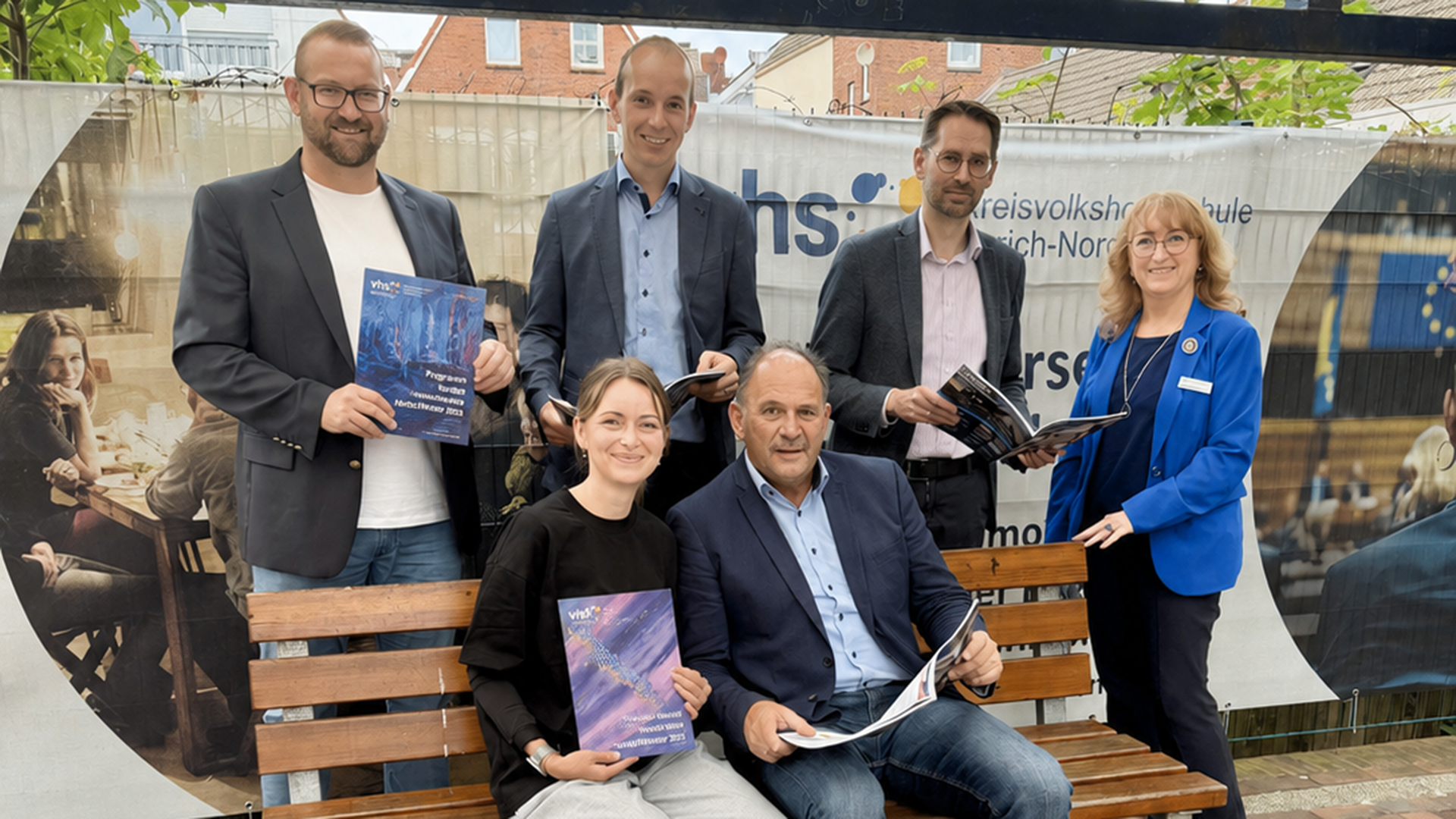 Gruppenfoto von sechs Personen vor einem Banner der Kreisvolkshochschule Aurich-Norden. Vier Männer und zwei Frauen stehen beziehungsweise sitzen um eine Holzbank und halten Programmhefte oder Broschüren in den Händen. Im Hintergrund sind das Logo und der Schriftzug der Kreisvolkshochschule zu sehen. Die Aufnahme entstand im Freien vor einer Kulisse mit Häusern und Grünpflanzen.