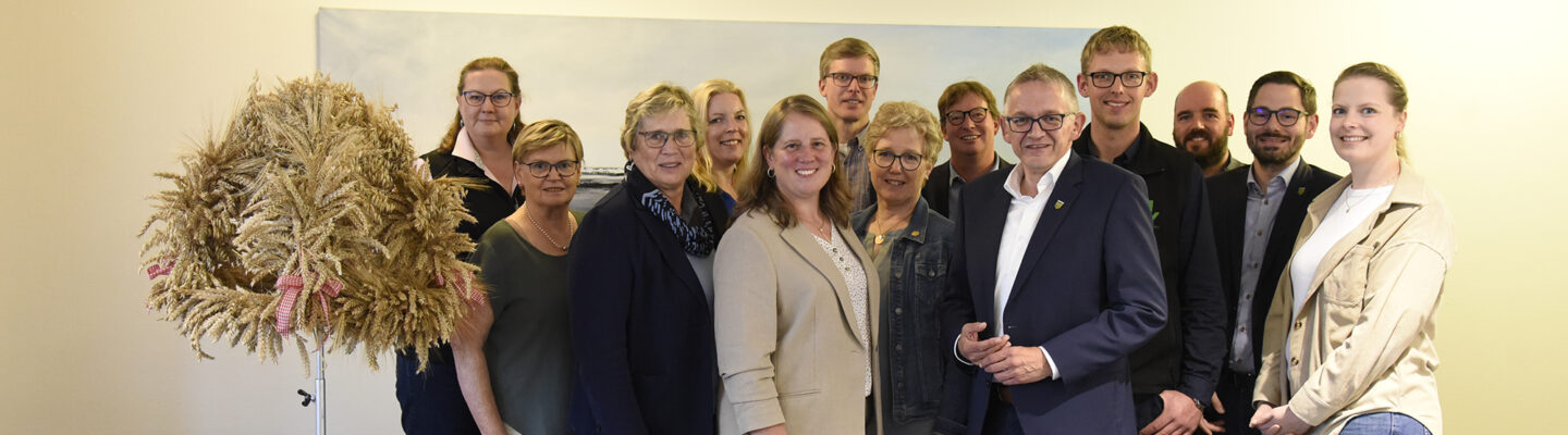Gruppenfoto von 13 Personen bei einer Veranstaltung des Landkreises Aurich. Die Gruppe steht lächelnd vor einer hellen Wand; links befindet sich eine große Erntekrone aus Getreideähren, daneben ein roter Sessel.