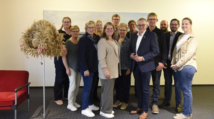 Gruppenfoto von 13 Personen bei einer Veranstaltung des Landkreises Aurich. Die Gruppe steht lächelnd vor einer hellen Wand; links befindet sich eine große Erntekrone aus Getreideähren, daneben ein roter Sessel.
