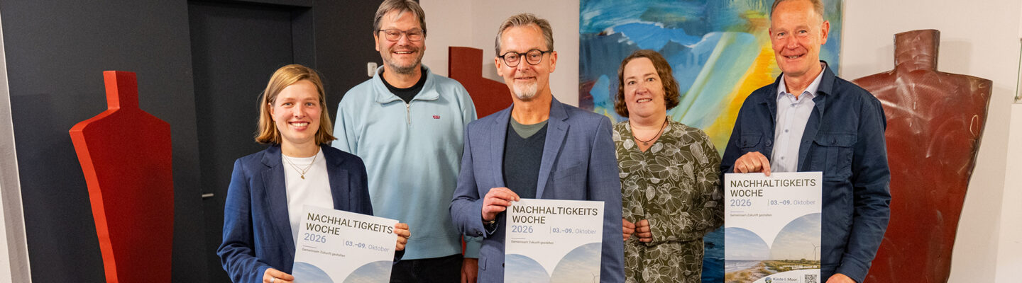 Fünf Personen stehen gemeinsam in einem Innenraum und präsentieren Plakate zur „Nachhaltigkeitswoche 2026“, die vom 3. bis 9. Oktober stattfindet. Im Hintergrund sind farbige Kunstwerke und große skulpturale Figuren zu sehen.