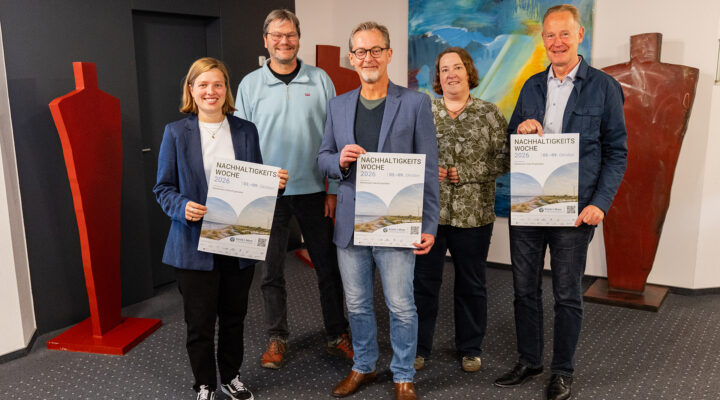 Fünf Personen stehen gemeinsam in einem Innenraum und präsentieren Plakate zur „Nachhaltigkeitswoche 2026“, die vom 3. bis 9. Oktober stattfindet. Im Hintergrund sind farbige Kunstwerke und große skulpturale Figuren zu sehen.