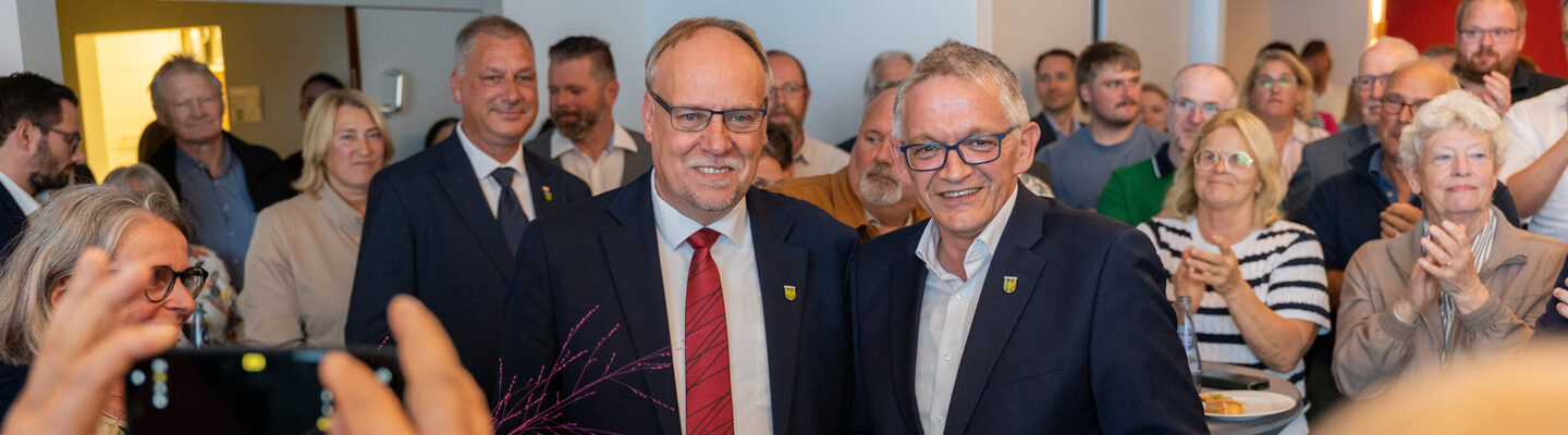 Zwei Männer in Anzügen stehen lächelnd nebeneinander inmitten einer größeren Menschenmenge. Im Vordergrund werden sie mit einem Smartphone und einer Kamera fotografiert, während mehrere Gäste im Hintergrund applaudieren.