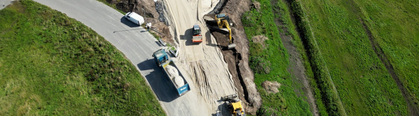 Luftaufnahme einer Straßenbaustelle mit Baggern, Radlader und Lkw. Eine neue Zufahrt wird auf einer Sandschicht zwischen angrenzenden Grünflächen angelegt.