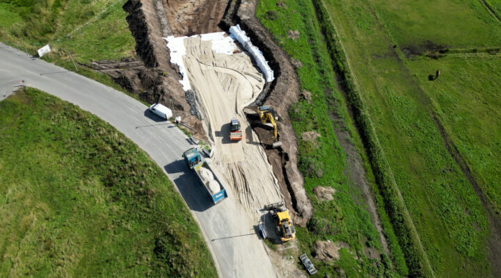 Luftaufnahme einer Straßenbaustelle mit Baggern, Radlader und Lkw. Eine neue Zufahrt wird auf einer Sandschicht zwischen angrenzenden Grünflächen angelegt.