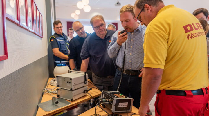 Mehrere Personen stehen bei einer Vorführung um technische Geräte auf Tischen. Ein Mitarbeiter der DLRG Wasserrettung bedient die Ausrüstung, während ein weiterer Mann in ein Funkgerät spricht und die übrigen Anwesenden die Demonstration verfolgen.