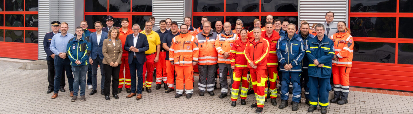Gruppenfoto von Einsatz- und Führungskräften verschiedener Hilfs- und Rettungsorganisationen vor der Feuerwehr-Technischen Zentrale des Landkreises Aurich. Die Gruppe steht vor mehreren großen roten Fahrzeugtoren; viele Personen tragen orange, rote oder blaue Einsatzkleidung.