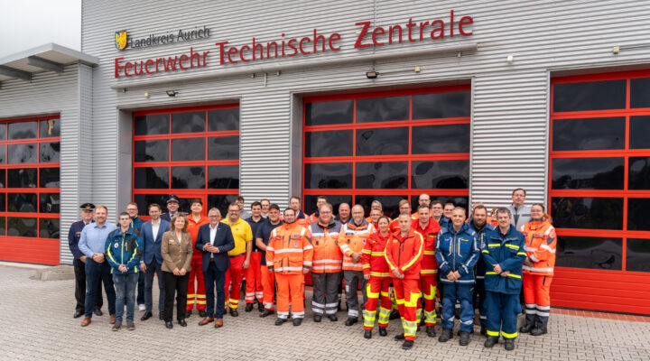 Gruppenfoto von Einsatz- und Führungskräften verschiedener Hilfs- und Rettungsorganisationen vor der Feuerwehr-Technischen Zentrale des Landkreises Aurich. Die Gruppe steht vor mehreren großen roten Fahrzeugtoren; viele Personen tragen orange, rote oder blaue Einsatzkleidung.