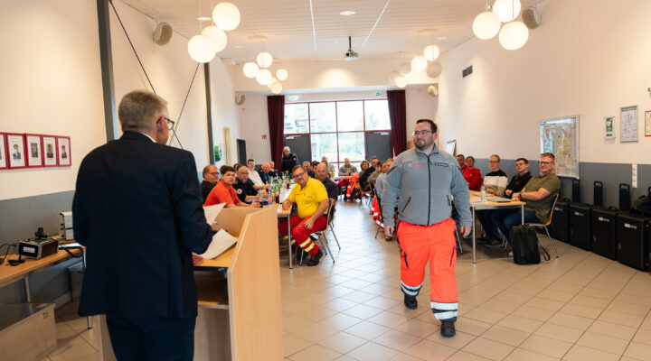 Blick in einen Schulungsraum der Feuerwehr-Technischen Zentrale: Zahlreiche Einsatzkräfte verschiedener Hilfsorganisationen sitzen an Tischen und verfolgen eine Veranstaltung. Im Vordergrund steht ein Mann in Einsatzkleidung im Mittelgang, während links ein Redner am Pult zu sehen ist.