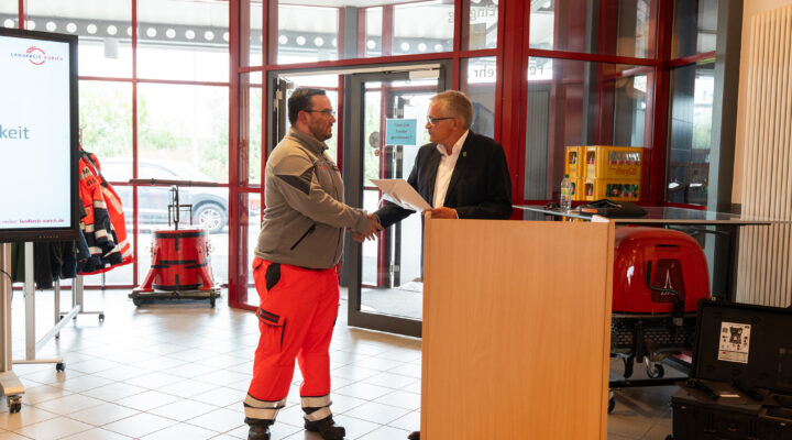 Ein Mitarbeiter einer Hilfsorganisation in grauer Jacke und orangefarbener Einsatzhose erhält bei einer Veranstaltung eine Urkunde und schüttelt dem Redner am Pult die Hand. Im Hintergrund sind Einsatzkleidung und technische Ausrüstung zu sehen.