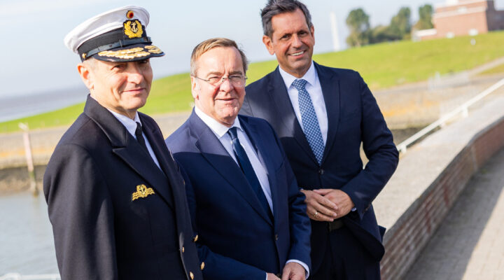 Drei Männer stehen bei sonnigem Wetter an einer Hafenanlage. Links trägt Vizeadmiral Wilhelm Tobias Abry eine Marineuniform mit weißer Schirmmütze, daneben stehen Verteidigungsminister Boris Pistorius und Niedersachsens Ministerpräsident Olaf Lies in dunklen Anzügen. Im Hintergrund sind ein Deich und eine maritime Anlage zu sehen.