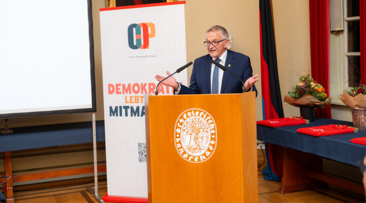 Landrat Olaf Meinen hält eine Begrüßungsrede bei einer Veranstaltung zum Carl-Osterwald-Preis. Er steht im dunklen Anzug hinter einem hölzernen Rednerpult mit dem Wappen des Landkreises Aurich und gestikuliert mit beiden Händen. Neben ihm steht ein Banner mit dem Logo des Carl-Osterwald-Preises und dem Schriftzug „Demokratie lebt vom Mitmachen“.