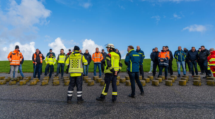 Zahlreiche Einsatzkräfte und Vertreter verschiedener Hilfsorganisationen stehen bei einer Einsatzbesprechung auf einem Deich. Im Vordergrund sind Mitglieder der Übungsleitung des Havariekommandos, der Feuerwehr und des Katastrophenschutzes in auffälliger Einsatzkleidung zu sehen. Im Hintergrund erstreckt sich eine grüne Deichlandschaft unter blauem Himmel. Die Aufnahme zeigt eine Besprechung im Rahmen einer Übung zur Chemikalienunfallbekämpfung an der Küste.