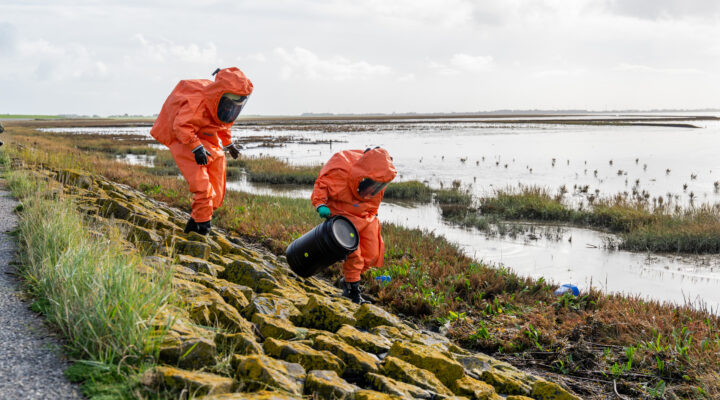 Zwei Einsatzkräfte in orangefarbenen Chemikalienschutzanzügen bewegen sich über eine steinige Uferbefestigung am Wattenmeer. Eine Person trägt ein schwarzes Fass, während die andere den Abstieg beobachtet. Im Hintergrund erstrecken sich Salzwiesen und Wattflächen unter bewölktem Himmel.