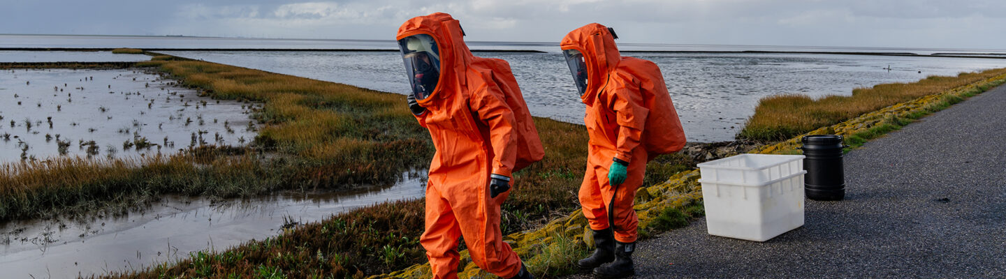 Zwei Einsatzkräfte in orangefarbenen Chemikalienschutzanzügen mit Atemschutzmasken bewegen sich entlang eines Deiches am Wattenmeer. Neben ihnen stehen eine weiße Kunststoffwanne und ein schwarzes Fass. Im Hintergrund erstrecken sich Salzwiesen und Wattflächen unter einem stark bewölkten Himmel.