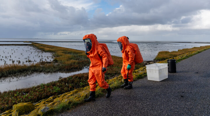 Zwei Einsatzkräfte in orangefarbenen Chemikalienschutzanzügen mit Atemschutzmasken bewegen sich entlang eines Deiches am Wattenmeer. Neben ihnen stehen eine weiße Kunststoffwanne und ein schwarzes Fass. Im Hintergrund erstrecken sich Salzwiesen und Wattflächen unter einem stark bewölkten Himmel.