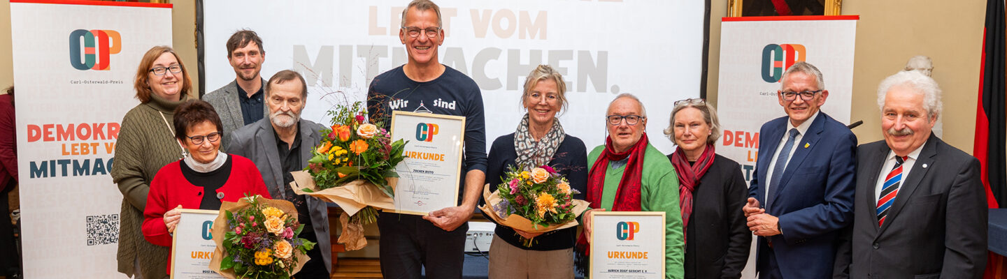Gruppenfoto der Preisträgerinnen und Preisträger des Carl-Osterwald-Preises 2026 gemeinsam mit weiteren Beteiligten der Preisverleihung. Mehrere Personen halten gerahmte Urkunden und bunte Blumensträuße in den Händen. Rechts stehen Landrat Olaf Meinen und ein weiterer Mann im Anzug. Im Hintergrund sind Banner des Carl-Osterwald-Preises mit dem Motto „Demokratie lebt vom Mitmachen“ zu sehen.