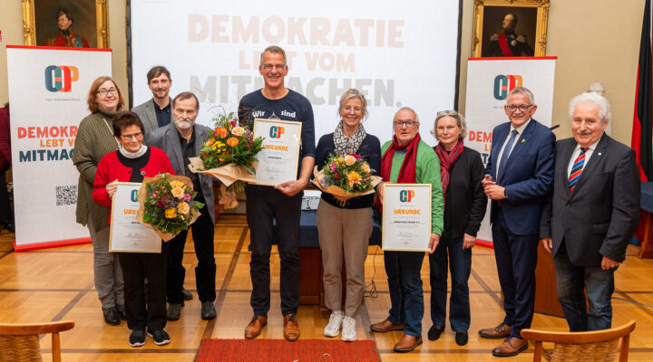 Gruppenfoto der Preisträgerinnen und Preisträger des Carl-Osterwald-Preises 2026 gemeinsam mit weiteren Beteiligten der Preisverleihung. Mehrere Personen halten gerahmte Urkunden und bunte Blumensträuße in den Händen. Rechts stehen Landrat Olaf Meinen und ein weiterer Mann im Anzug. Im Hintergrund sind Banner des Carl-Osterwald-Preises mit dem Motto „Demokratie lebt vom Mitmachen“ zu sehen.