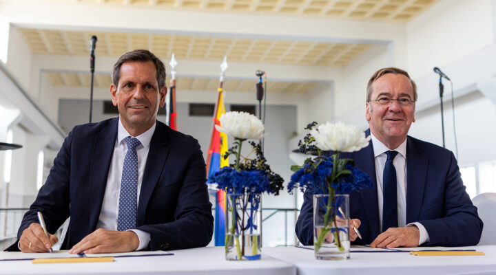 Niedersachsens Ministerpräsident Olaf Lies und Bundesverteidigungsminister Boris Pistorius sitzen an zwei Stehtischen und unterzeichnen Dokumente. Zwischen ihnen stehen Blumenvasen mit weißen und blauen Blüten; im Hintergrund sind Fahnen und Mikrofone zu sehen.