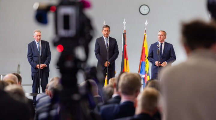 Drei Männer in dunklen Anzügen stehen bei einer Veranstaltung an Mikrofonen vor mehreren Fahnen. In der Mitte steht Niedersachsens Ministerpräsident Olaf Lies, rechts Bundesverteidigungsminister Boris Pistorius. Im Vordergrund sind das sitzende Publikum sowie unscharf eine Fernsehkamera zu sehen.
