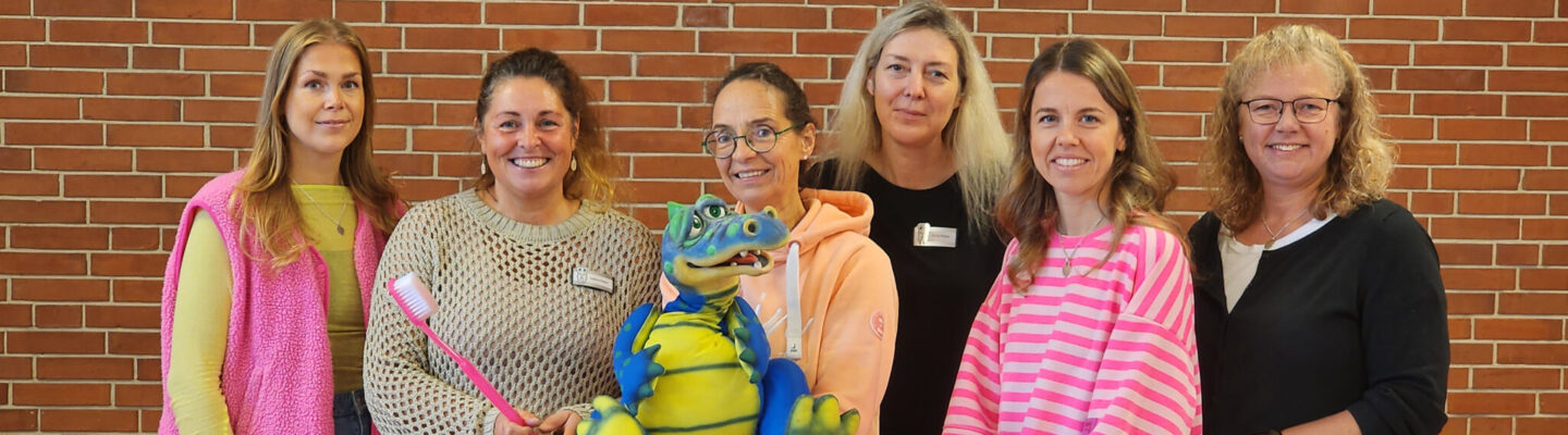Sechs Frauen stehen vor einer roten Backsteinwand und lächeln in die Kamera. In ihrer Mitte befindet sich eine bunte Krokodilfigur neben einer übergroßen Zahnbürste und einem Modell, das an Zähne beziehungsweise Zahnpflege erinnert.