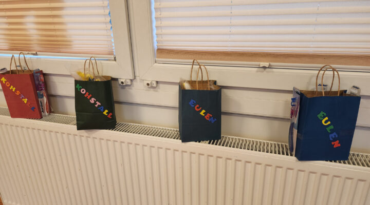 Vier farbige Papiertüten stehen auf einer Fensterbank über einem Heizkörper. Sie sind mit bunten Buchstaben beschriftet, zweimal mit „KOHSTALL“ und zweimal mit „EULEN“, und enthalten unter anderem verpackte Zahnbürsten und weitere kleine Gegenstände.