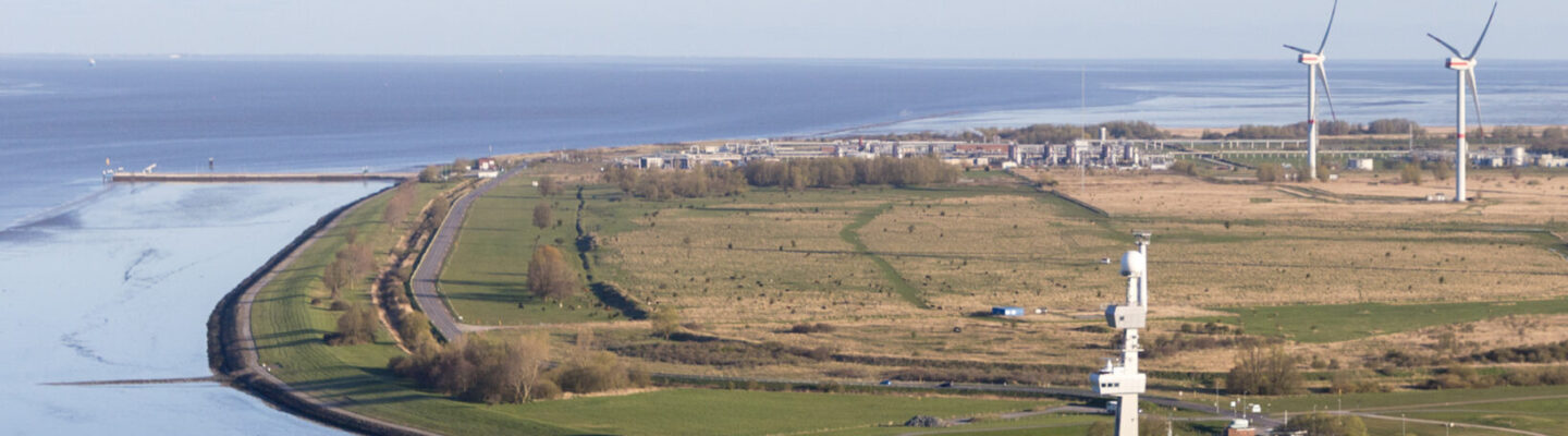 Luftaufnahme einer flachen Küstenlandschaft an der Nordsee mit Deichen, Wasserflächen und Grünland. Nahe der Küste steht eine hohe maritime Anlage; im Hintergrund sind zwei Windenergieanlagen sowie Industrieanlagen zu sehen.
