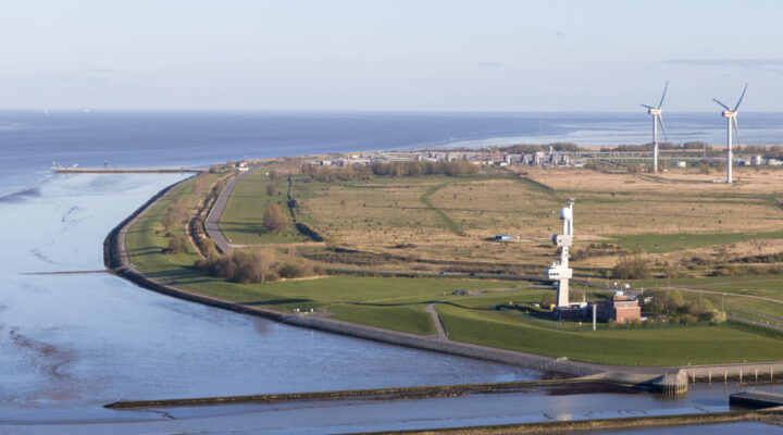 Luftaufnahme einer flachen Küstenlandschaft an der Nordsee mit Deichen, Wasserflächen und Grünland. Nahe der Küste steht eine hohe maritime Anlage; im Hintergrund sind zwei Windenergieanlagen sowie Industrieanlagen zu sehen.