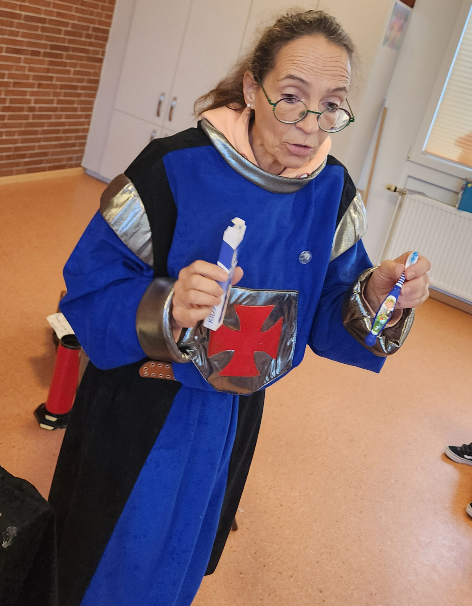 Eine Frau in einem blau-schwarzen, mittelalterlich anmutenden Kostüm mit silberfarbenen Verzierungen und rotem Kreuz hält eine verpackte sowie eine Kinderzahnbürste in den Händen und scheint diese zu erklären oder vorzuführen.