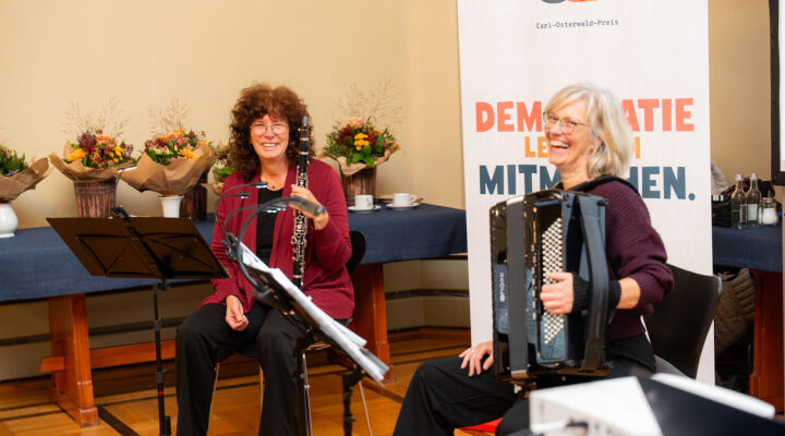 Zwei Musikerinnen der Musikschule Landkreis Aurich, Frau Anja Lütke-Notarp (Klarinette) und Frau Edda Liebermann-Pauen (Akkordeon), gestalten die musikalische Umrahmung der Verleihung des Carl-Osterwald-Preises. Die Frauen sitzen lächelnd mit einer Klarinette und einem Akkordeon vor Notenständern. Im Hintergrund stehen Blumensträuße und ein Banner mit dem Motto „Demokratie lebt vom Mitmachen“.