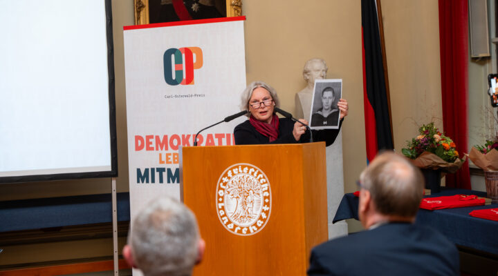 Hilke Osterwald hält bei der Verleihung des Carl-Osterwald-Preises 2026 eine Rede hinter einem hölzernen Rednerpult mit dem Wappen des Landkreises Aurich. Dabei zeigt sie dem Publikum ein historisches Schwarz-Weiß-Foto ihres Vaters Carl Osterwald im Alter von 17 Jahren, auf dem er eine Matrosenuniform trägt. Im Hintergrund steht ein Banner des Carl-Osterwald-Preises mit dem Motto „Demokratie lebt vom Mitmachen“.