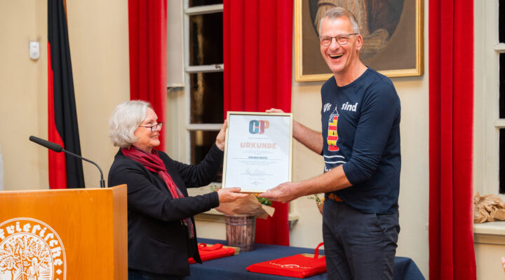 Hilke Osterwald überreicht Jochen Risto aus der Krummhörn die Urkunde des Carl-Osterwald-Preises 2026. Beide halten die gerahmte Urkunde gemeinsam in den Händen und lächeln sich an. Jochen Risto trägt ein dunkelblaues Langarmshirt mit Leuchtturmmotiv. Im Hintergrund sind rote Vorhänge, eine Deutschlandflagge und ein Tisch mit roten Geschenktaschen zu sehen.